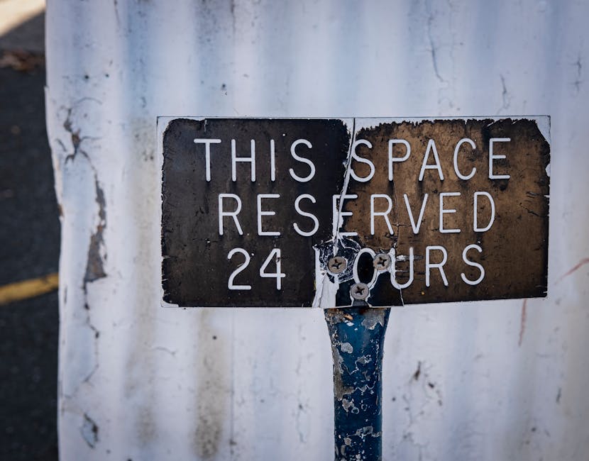 A close-up photograph of a weathered metal sign mounted on a blue pole, attached to a white painted wall. The sign reads 'THIS SPACE RESERVED 24 HOURS' in white capital letters, with some paint chipping and rust visible on its surface, indicating age and exposure to the elements. The background includes a section of asphalt pavement with a yellow line, suggesting an outdoor parking or loading area. The image emphasizes the sign's worn condition and surroundings, providing context for vehicle access and scheduling restrictions relevant to house removals and moving logistics managed by Man with Van Upper Norwood.