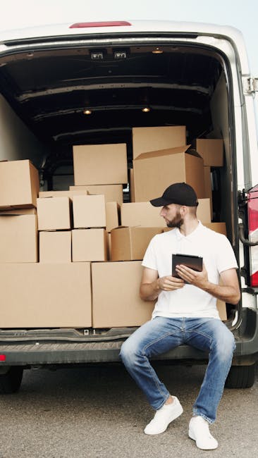 A man wearing a white t-shirt, blue jeans, and a black cap is seated on the edge of an open van’s rear door, which is filled with stacked cardboard boxes of varying sizes, some sealed with packing tape. He is holding a tablet device and appears to be reviewing or managing the packing and loading process during a home relocation or furniture transport. The van is parked outdoors on a paved surface, with a dark interior visible into the vehicle’s cargo space. The boxes are arranged systematically to optimize space for moving items such as household belongings, and the scene emphasizes professional removals services provided by Man with Van Upper Norwood. The environment suggests an organized loading and packing operation typical of house removals, with materials like cardboard and packing tape evident in the cargo area.