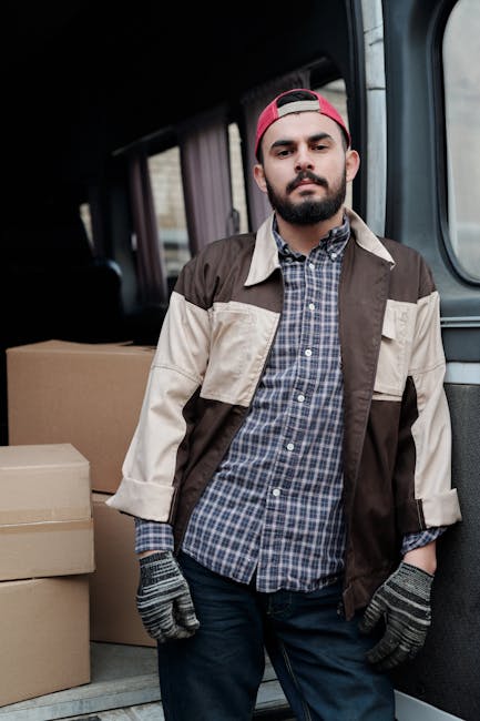 A man with a beard, wearing a red baseball cap backwards, a checkered shirt, and a beige and brown jacket, stands inside the open rear of a van used for house removals. Inside the vehicle, several cardboard boxes are stacked, some wrapped in plastic, ready for transport. The man is wearing gloves and appears to be involved in a home relocation process, with the van positioned on a paved area near a building entrance. The scene depicts the loading or unloading stage of furniture transport and packing during a professional removals service provided by Man with Van Upper Norwood, supporting efficient move management in the neighborhood of Upper Norwood SE19.