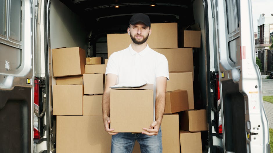 A man wearing a white t-shirt, blue jeans, and a black cap is seated on the edge of an open van’s rear door, which is filled with stacked cardboard boxes of varying sizes, some sealed with packing tape. He is holding a tablet device and appears to be reviewing or managing the packing and loading process during a home relocation or furniture transport. The van is parked outdoors on a paved surface, with a dark interior visible into the vehicle’s cargo space. The boxes are arranged systematically to optimize space for moving items such as household belongings, and the scene emphasizes professional removals services provided by Man with Van Upper Norwood. The environment suggests an organized loading and packing operation typical of house removals, with materials like cardboard and packing tape evident in the cargo area.