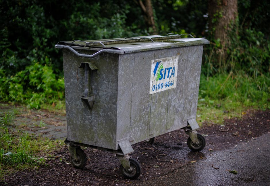 A large, weathered grey waste disposal bin with a partially open lid, positioned on a paved surface outside a residential property in an outdoor setting with green foliage and trees in the background. The bin has four black caster wheels, with two visible at the front and one at the rear, and features a white and blue SITA logo along with a contact number. The surrounding ground appears damp, with some mud and small debris, indicating recent clearing or loading activities related to home relocation or waste clearance. This scene captures the loading process of such bins for collection as part of a house removal or packing and moving service by Man with Van Upper Norwood, supporting proper disposal practices in accordance with Bromley Council's skip and disposal rules for SE19, UPPER NORWOOD.