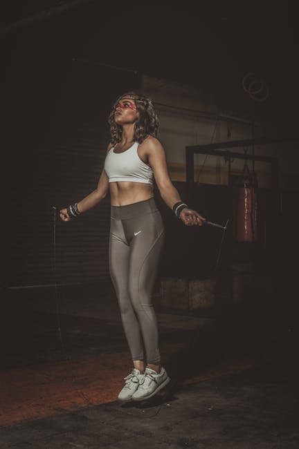 A woman with curly hair, dressed in a white sports bra and grey Nike leggings, is captured mid-jump while skipping with a jump rope inside a gym or fitness studio. She appears focused and energetic, with her arms extended holding the jump rope handles and her feet slightly off the ground. The environment has a dark background with gym equipment such as a punching bag hanging on the wall and a floor mat visible beneath her. The lighting emphasizes her athletic outfit and active pose. This image relates to home or personal fitness routines, which can be part of a healthy lifestyle during house removals or relocation preparations and can be associated with professional moving or fitness services provided by companies like Man with Van Upper Norwood.