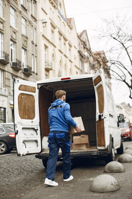 A man wearing a blue hoodie and blue trousers is standing on a cobblestone street next to an open white cargo van used for house removals, with the van positioned close to a historic urban building wi