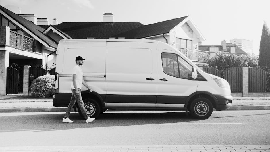 A man wearing a blue hoodie and blue trousers is standing on a cobblestone street next to an open white cargo van used for house removals, with the van positioned close to a historic urban building wi