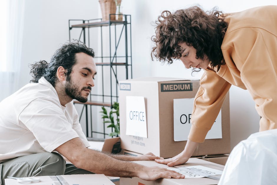 Two individuals are engaged in packing and organizing office belongings during a home relocation process within an indoor setting. The man with dark curly hair and a beard, dressed in a white shirt, is seated on the floor, carefully handling a cardboard box labeled 'OFFICE'. The woman, with voluminous curly hair and wearing a beige jacket, is leaning over and pressing down on a document or packing material on top of the box. Surrounding them are several packed moving boxes, some labeled with small signs indicating the contents or size, and a few open containers with packing materials such as plastic wrap and paper. In the background, a metal shelving unit and a potted plant are visible, suggesting a residential or small office environment. The scene illustrates the detailed packing stage of a furniture transport and office relocation, with visible attention to organized labeling, secure packing, and the physical handling of items. Man with Van Upper Norwood's professional moving team is engaged in the packing and moving process at this indoor location, supporting small business relocations or home-based office moves.