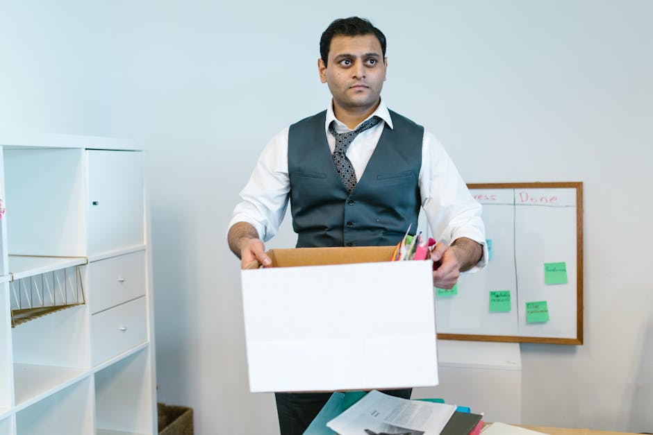 A man with dark hair, wearing a white dress shirt, a dark grey vest, and a patterned tie, is standing indoors holding an open cardboard box filled with pens, pencils, and other office stationery. Behind him, there is a whiteboard with green sticky notes and handwritten notes, and to his left, a white storage unit with multiple compartments. The room appears to be an office environment with neutral lighting and minimal decor. The man is engaged in a packing or moving process typical of office relocation services, representing furniture transport, packing, and logistical activities associated with professional removals. This image is related to house and office removals services offered by Man with Van Upper Norwood, supporting the context of office relocation as part of moving and transport logistics.