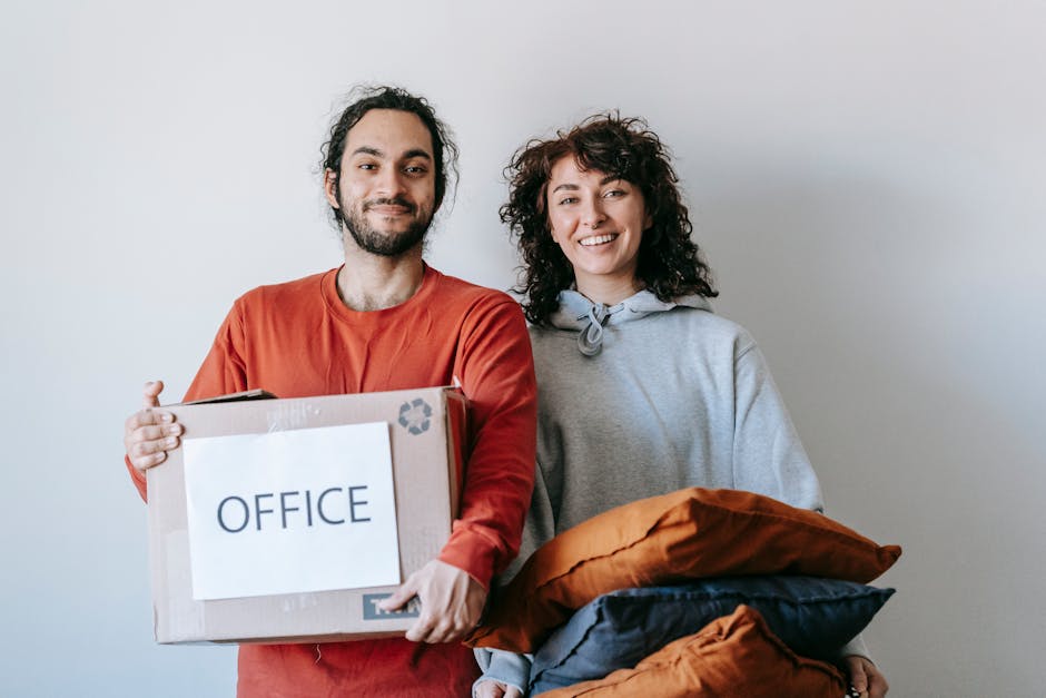 Two individuals are engaged in packing and organizing office belongings during a home relocation process within an indoor setting. The man with dark curly hair and a beard, dressed in a white shirt, is seated on the floor, carefully handling a cardboard box labeled 'OFFICE'. The woman, with voluminous curly hair and wearing a beige jacket, is leaning over and pressing down on a document or packing material on top of the box. Surrounding them are several packed moving boxes, some labeled with small signs indicating the contents or size, and a few open containers with packing materials such as plastic wrap and paper. In the background, a metal shelving unit and a potted plant are visible, suggesting a residential or small office environment. The scene illustrates the detailed packing stage of a furniture transport and office relocation, with visible attention to organized labeling, secure packing, and the physical handling of items. Man with Van Upper Norwood's professional moving team is engaged in the packing and moving process at this indoor location, supporting small business relocations or home-based office moves.