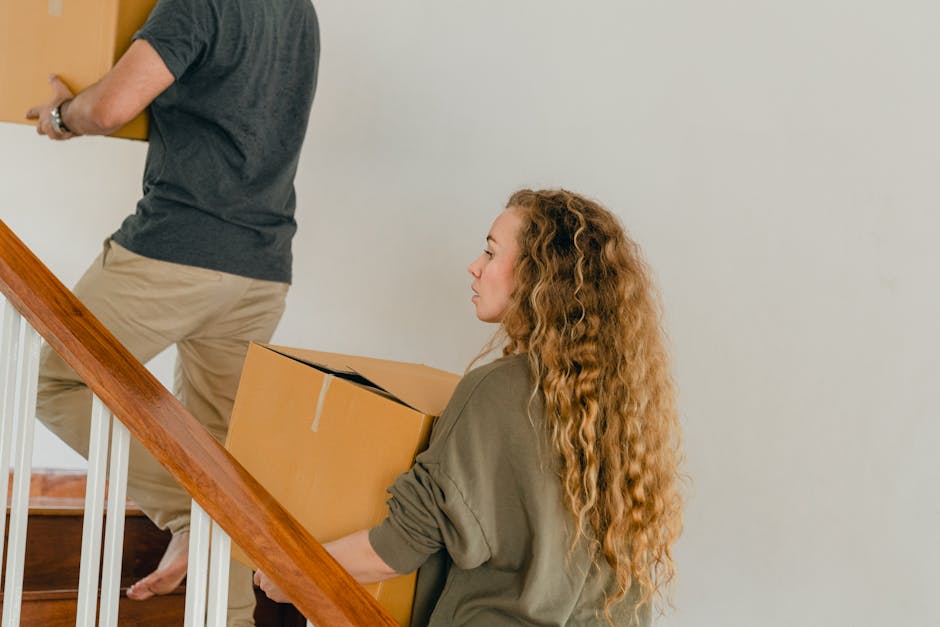 A woman with long, curly red hair carrying a medium-sized cardboard box while ascending a wooden staircase inside a home, with a man in casual clothing partially visible ahead of her holding a similar box, during a home relocation process. The staircase has a wooden handrail and white spindles, with the interior space illuminated by natural light. Behind them, the plain white wall and wooden flooring are visible, indicating an organized packing and moving activity. The scene exemplifies furniture transport and packing procedures associated with professional removals services provided by Man with Van Upper Norwood, supporting efficient home relocation and moving logistics on Crown Lane, Upper Norwood.