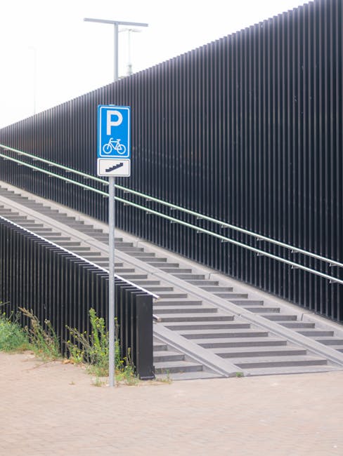 A pedestrian ramp with metal steps and handrails leading up to an elevated area, positioned beside a tall, vertical, black metal fence. A blue parking sign indicating bicycle and vehicle parking is mounted on a pole near the ramp, situated on a paved surface with some small green plants and grass at the base. The scene is outdoor, with bright natural lighting. This image relates to house removals and moving logistics as part of the process of loading and transporting belongings during a home relocation. The sign and ramp are situated on Crown Lane in Upper Norwood, supporting the context of traffic and parking considerations for professional moving services by Man with Van Upper Norwood, who specialise in furniture transport and packing for house moves.