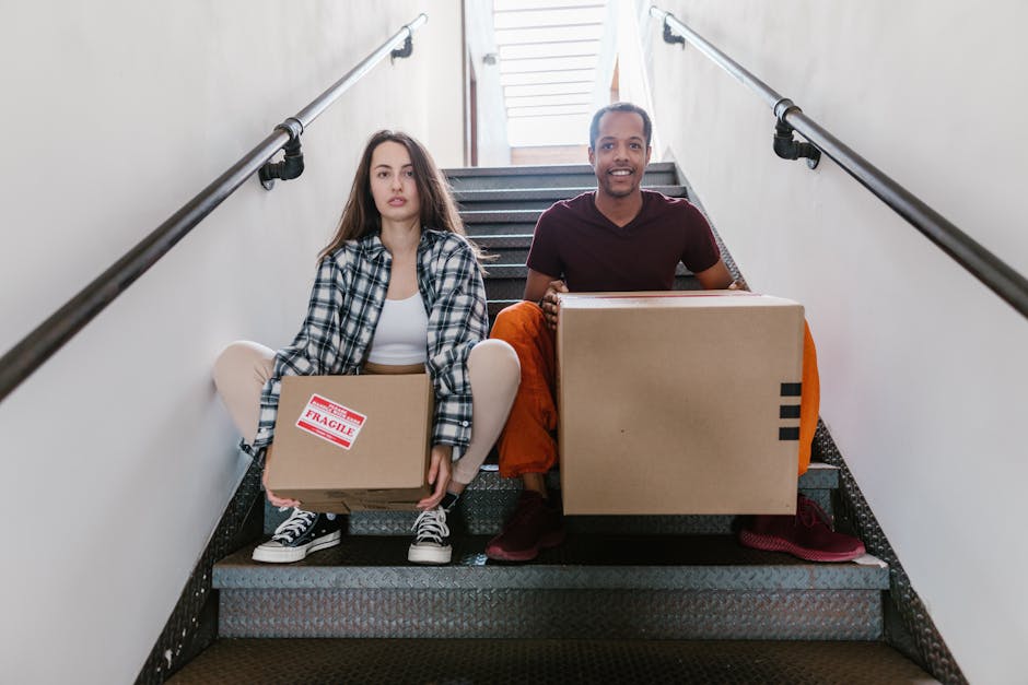A woman with long, curly red hair carrying a medium-sized cardboard box while ascending a wooden staircase inside a home, with a man in casual clothing partially visible ahead of her holding a similar box, during a home relocation process. The staircase has a wooden handrail and white spindles, with the interior space illuminated by natural light. Behind them, the plain white wall and wooden flooring are visible, indicating an organized packing and moving activity. The scene exemplifies furniture transport and packing procedures associated with professional removals services provided by Man with Van Upper Norwood, supporting efficient home relocation and moving logistics on Crown Lane, Upper Norwood.