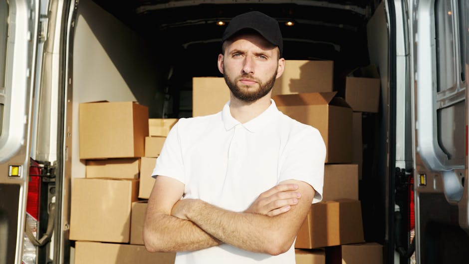 A man wearing a white polo shirt and a black cap stands with arms crossed inside the rear of a moving van loaded with multiple cardboard boxes of various sizes, some wrapped in plastic or fabric for protection, arranged on the floor and against the walls of the van. The van's interior is illuminated, revealing the wooden flooring and metal sides, and the boxes appear prepared for home relocation or furniture transport as part of a professional house removal service. Outside the van, at the doorway, the pavement and partial view of the vehicle's exterior are visible, indicating an active loading process associated with packing and moving activities on a residential property in Upper Norwood, SE19. The overall scene reflects the careful handling and organization of household items during a furniture transport operation, with a focus on safe loading procedures, as provided by Man with Van Upper Norwood.