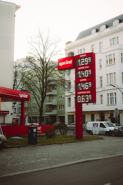 An outdoor scene featuring a tall red sign displaying fuel prices at a petrol station, with diesel priced at £1.299, Super E10 at £1.409, Super at £1.469, and Autogas LPG at £0.639. The sign is situated on a paved area near a sidewalk, with a leafless tree and residential buildings visible in the background. A white van is parked adjacent to the station, and a black trash bin is positioned nearby. On the left side, part of a red and black fuel pump canopy with the 'Sprint' logo can be seen, indicating it's a fuel station. This image relates to house removals and moving services provided by Man with Van Upper Norwood, illustrating the typical urban environment where such relocation services operate, possibly in support of home relocation or furniture transport involving careful planning and logistics.
