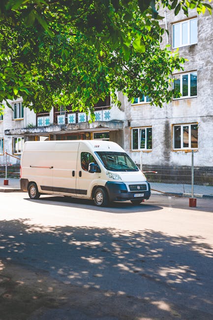 A white delivery van with a streamlined body and black front bumper is parked on a paved street adjacent to a residential building, partly shaded by large green tree leaves in the foreground. The building behind the van is an upper Norwood apartment with several windows, some with open curtains, and a weathered, concrete exterior. The van is positioned next to a sidewalk, and the area appears quiet and suitable for home relocation tasks. The surrounding environment includes safety barriers along the pavement, and the vehicle is aligned parallel to the curb, possibly involved in furniture transport or packing and moving procedures as part of a professional removals service by Man with Van Upper Norwood.