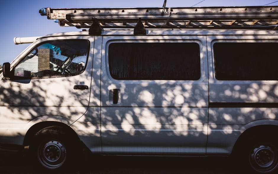 A white cargo van parked on a paved driveway outside a residential property during daylight. The van's side door is closed, and it features three windows along the side, with a large metal roof rack carrying a long, wooden ladder secured with straps. Shade and dappled sunlight from nearby trees cast shadows across the van’s exterior. The van is positioned next to the entrance of a house, indicating its involvement in house removals or furniture transport. The background includes the front of the house with a brick wall and a partially visible window. Inside the property, there appears to be packing materials such as cardboard boxes and wrapped furniture, suggesting ongoing packing or home relocation activities. The image decorates the context of professional removals services provided by Man with Van Upper Norwood, specializing in house and furniture movers, with the vehicle ready for loading or unloading furniture and household items during a home move on Sylvan Hill.