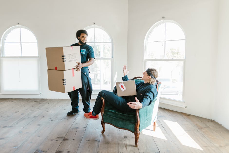 A man with medium skin tone and curly black hair sits on a wooden floor inside a room with large, arched windows that allow natural daylight to illuminate the space. He is dressed in a blue T-shirt and dark pants, and appears to be resting or waiting amidst several cardboard moving boxes of varying sizes, some of which have red packing tape. The boxes are stacked or placed nearby, indicating the process of packing or unpacking during a home relocation. A tall potted plant with broad green leaves is positioned against the wall next to the windows. The room has a minimalist appearance with clean white walls, and the natural light from outside highlights the details of the scene. This setting reflects the interior environment during a furniture transport or packing and moving operation, and illustrates part of the logistics involved in a house removal, a service offered by Man with Van Upper Norwood.